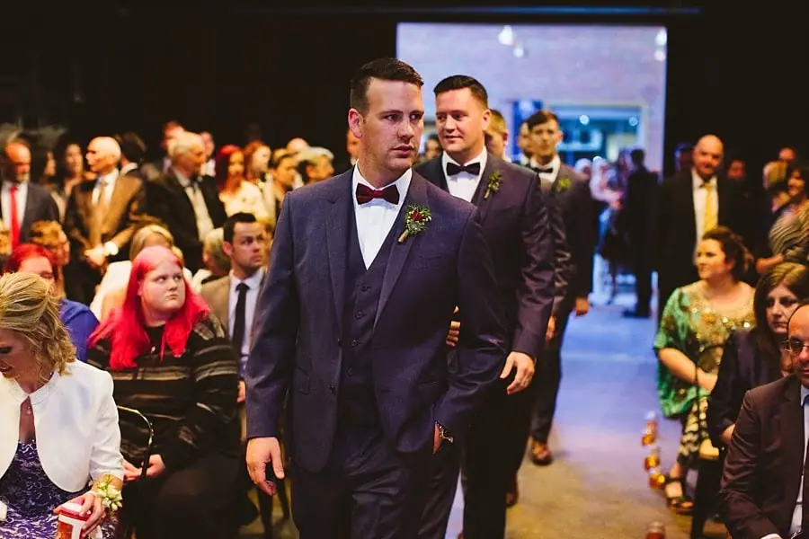 Groom walking past seated guests along an indoor ceremony aisle.