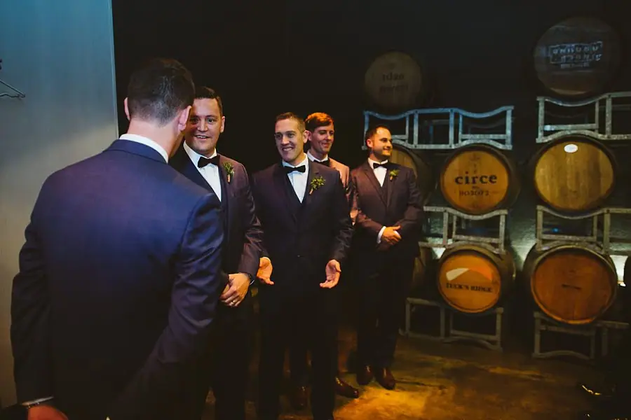 Groom standing with suited men beside stacked wine barrels.