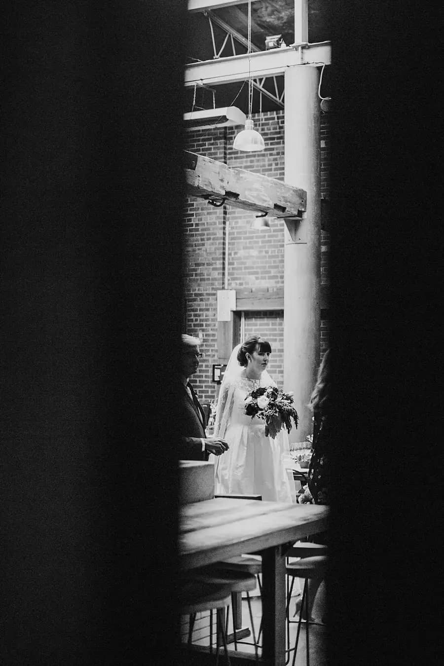 Black-and-white photograph of the bride visible through a narrow opening in a doorway.