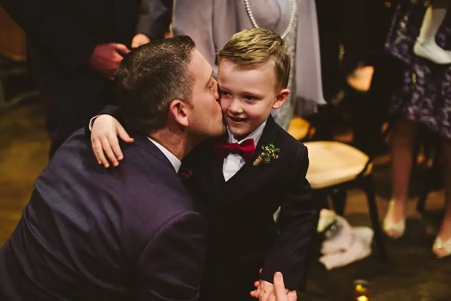 Groom kissing a smiling boy wearing a suit and bow tie.