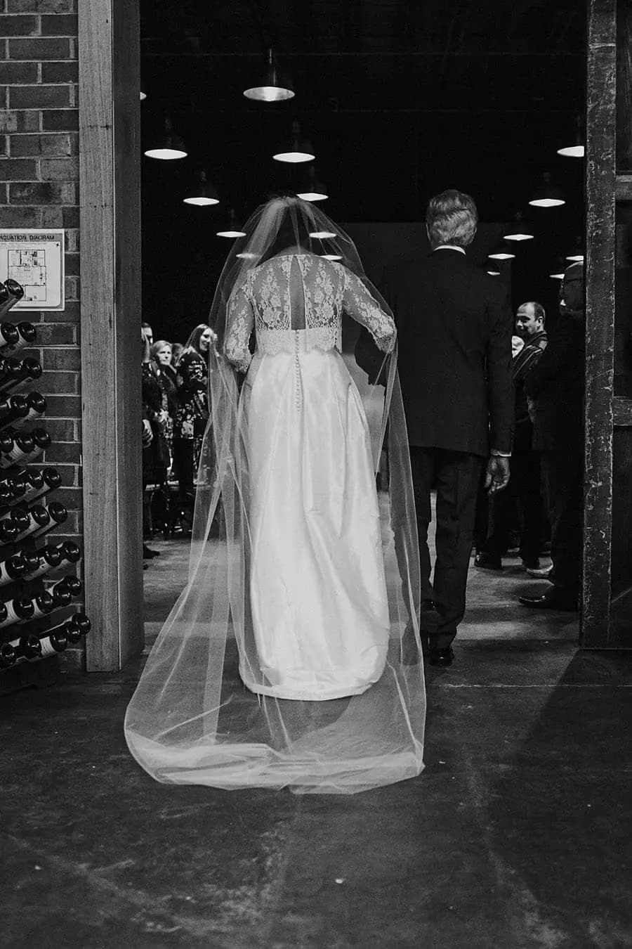 Black-and-white photograph of the veiled bride entering through a doorway with a suited man.