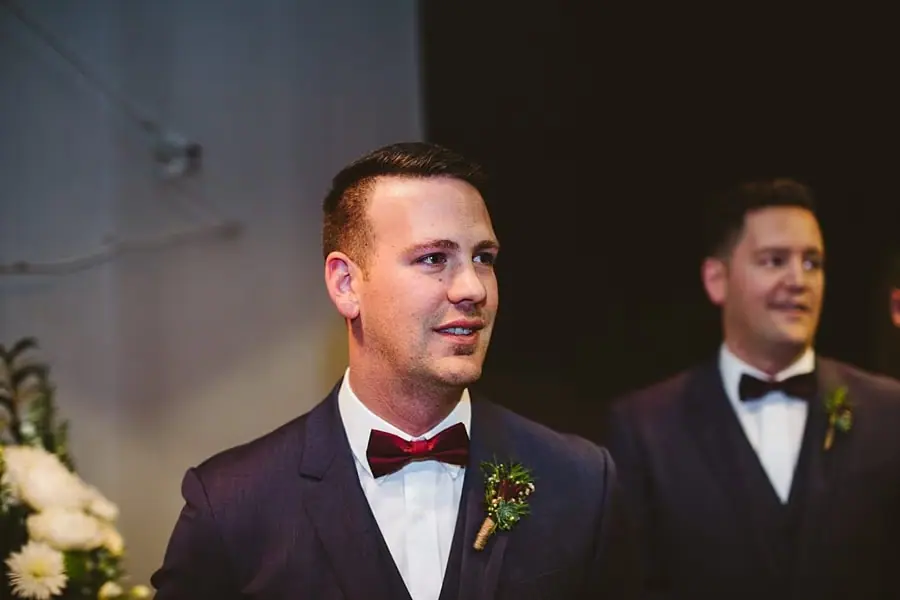 Groom watching the indoor wedding ceremony beside another suited man.