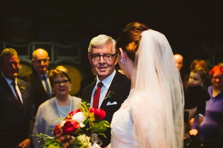 Older suited man smiling at the bride as they stand among ceremony guests.