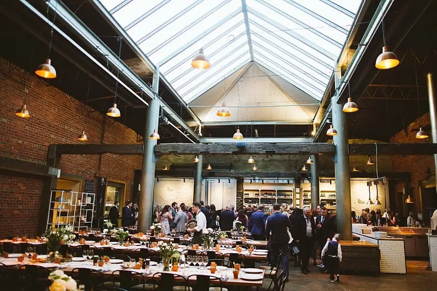 Guests gathering among reception tables beneath a large glass roof in a brick-walled room.