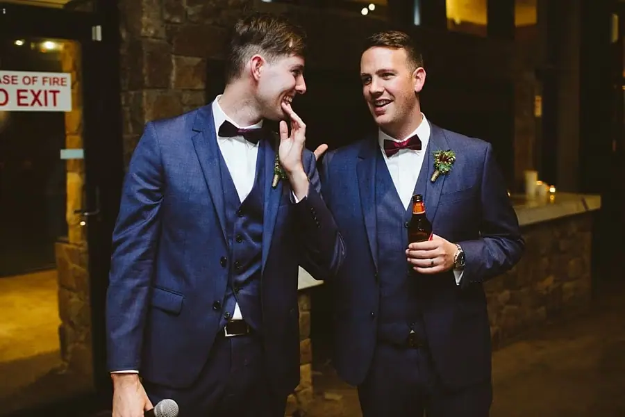 Two men in blue suits laughing together, one holding a microphone and the other a drink.