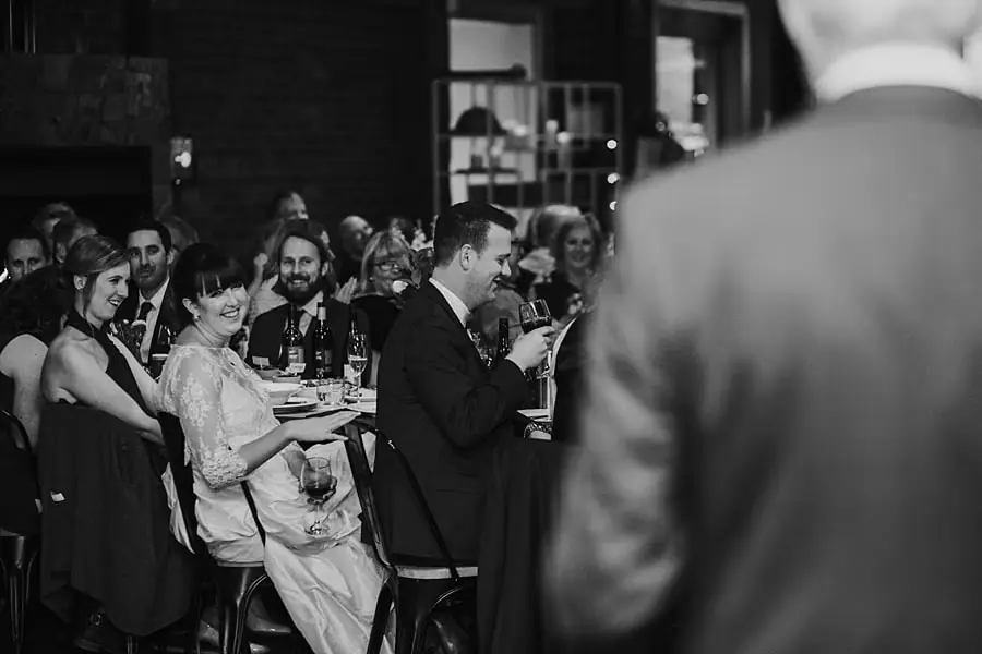 Black-and-white photograph of the bride and seated guests listening to a reception speech.