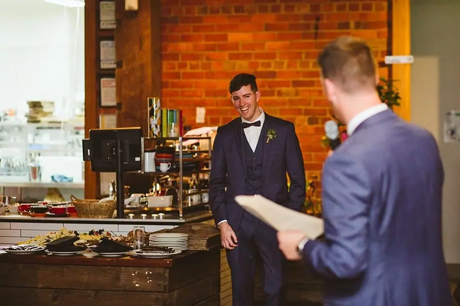 Man in a blue suit giving a speech while another suited man stands beside a reception table.