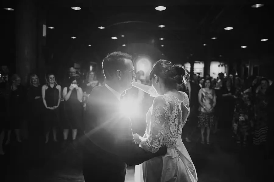 Black-and-white photograph of the couple dancing in front of watching guests and a bright light.