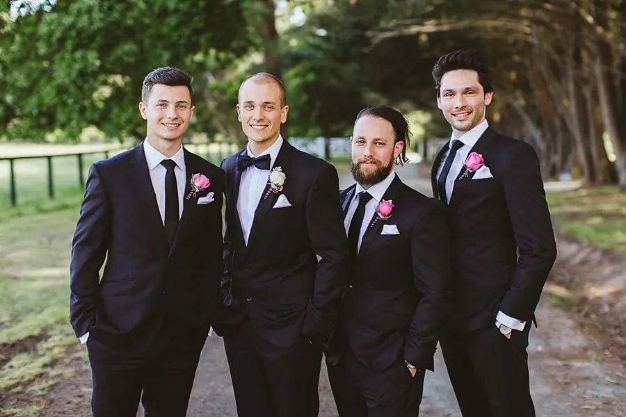 Four men in dark suits posing together beneath trees, wearing pink boutonnieres.