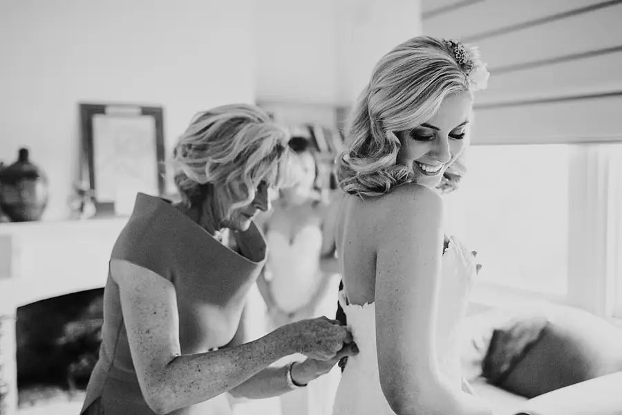 Black-and-white photograph of a woman helping fasten the back of the bride’s dress.