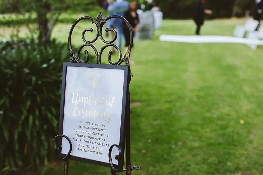 Framed wedding welcome sign on a metal stand beside a garden path.
