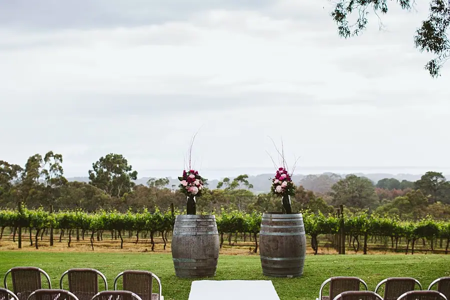 Outdoor ceremony area with rows of chairs and flower arrangements on wooden barrels overlooking vines.