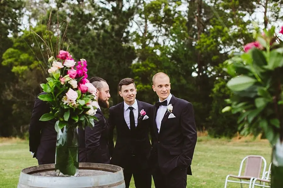 Two men in dark suits standing beside a tall floral arrangement before the ceremony.