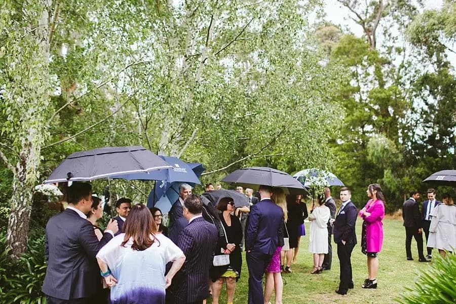 Wedding guests gathering under umbrellas on the lawn before the ceremony.