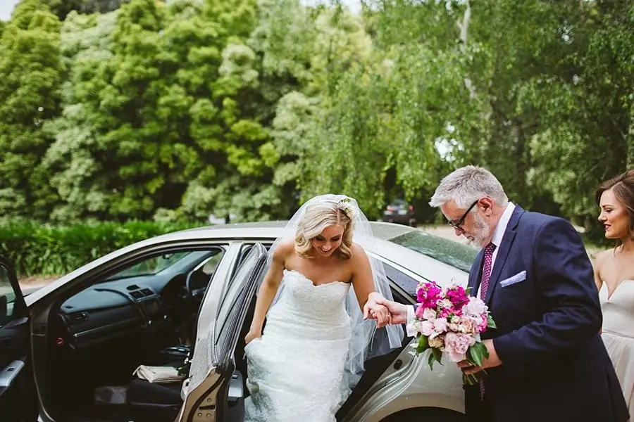 Bride stepping out of a car with help from a suited man while holding her bouquet.