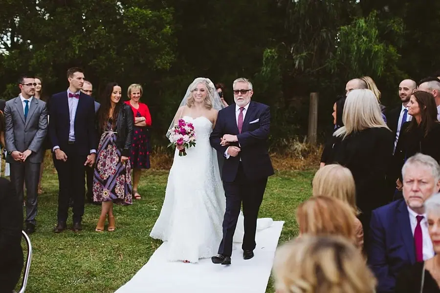 Bride walking down a white aisle runner on the arm of a man in a dark suit.