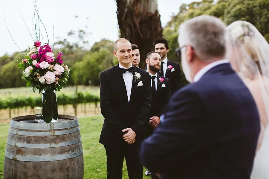 Groom smiling as the bride approaches the ceremony with a suited man.