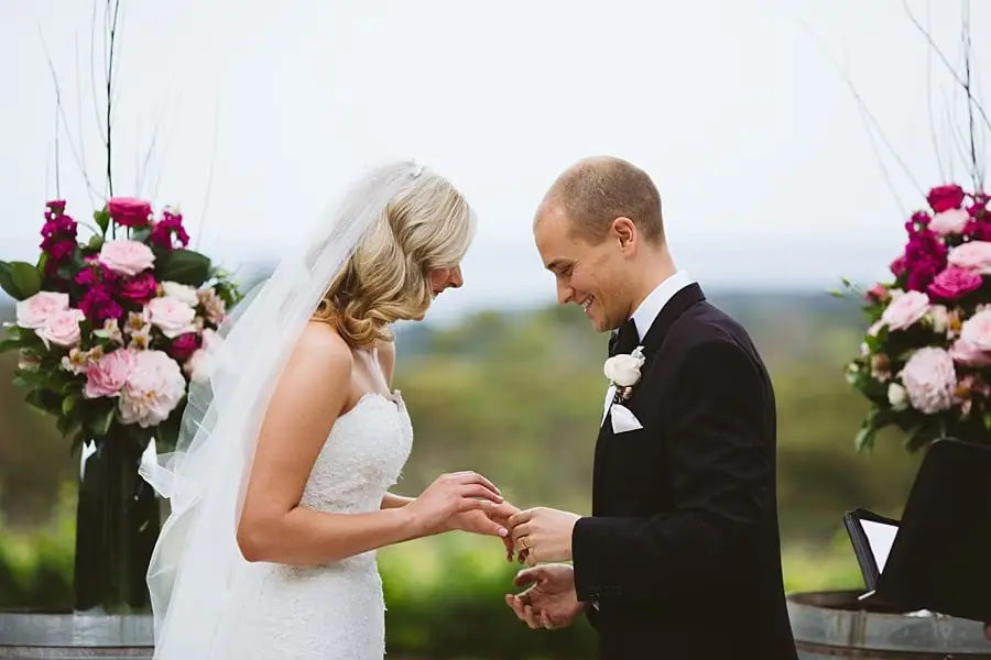 Bride and groom looking down at their joined hands beside pink floral arrangements.