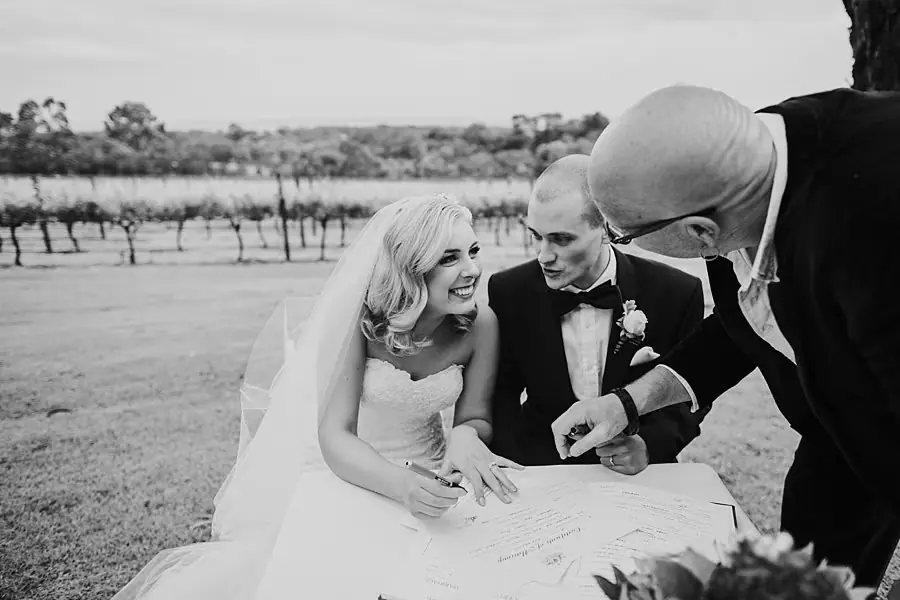 Black-and-white photograph of newlyweds signing a document at a table with their celebrant beside them.