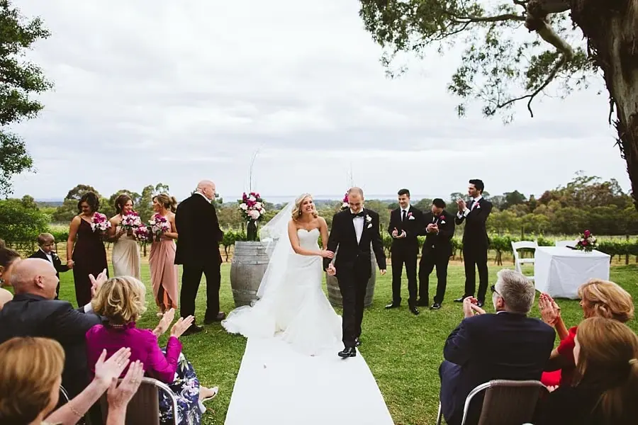 Newlyweds walking down the white aisle runner as seated guests applaud.