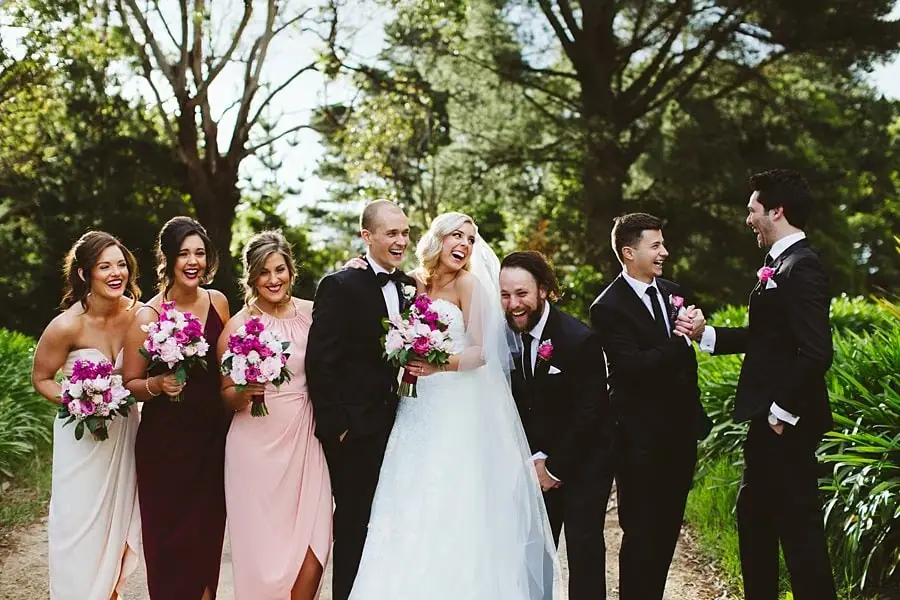 Bride, groom and wedding party laughing together beneath leafy trees.