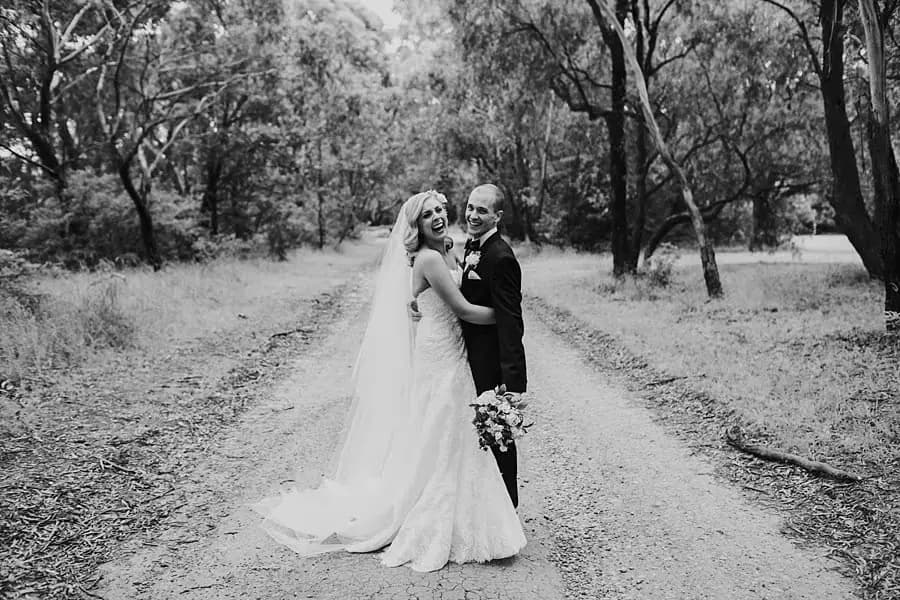 Black-and-white portrait of newlyweds embracing on a gravel path beneath trees.