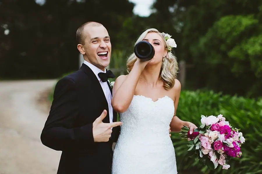 Groom smiling as the bride drinks from a dark bottle beside him on the path.