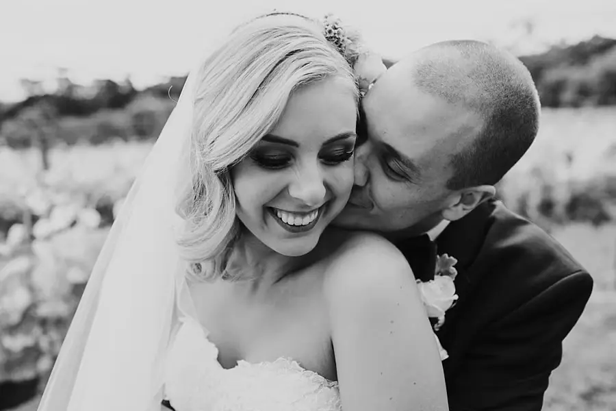 Black-and-white close portrait of the groom kissing the bride’s cheek.