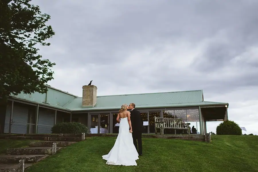 Bride and groom embracing on a lawn in front of a low reception building.