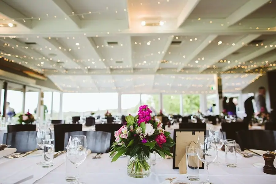 Pink flowers on a reception table beneath strings of ceiling lights in a bright dining room.