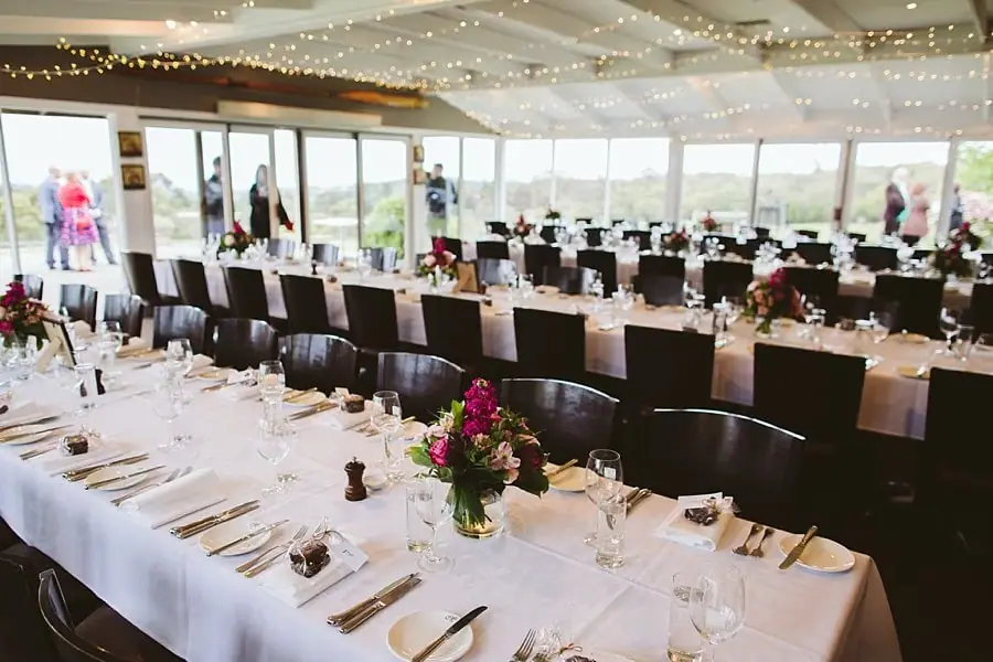 Long reception tables set with white linen, flowers, glasses and dark chairs beside large windows.