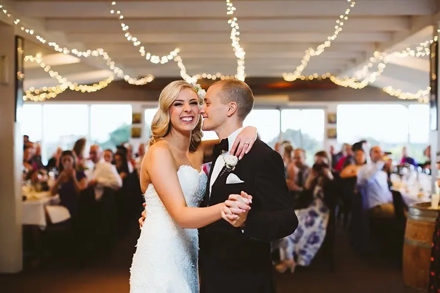 Bride and groom dancing together while reception guests look on beneath strings of lights.