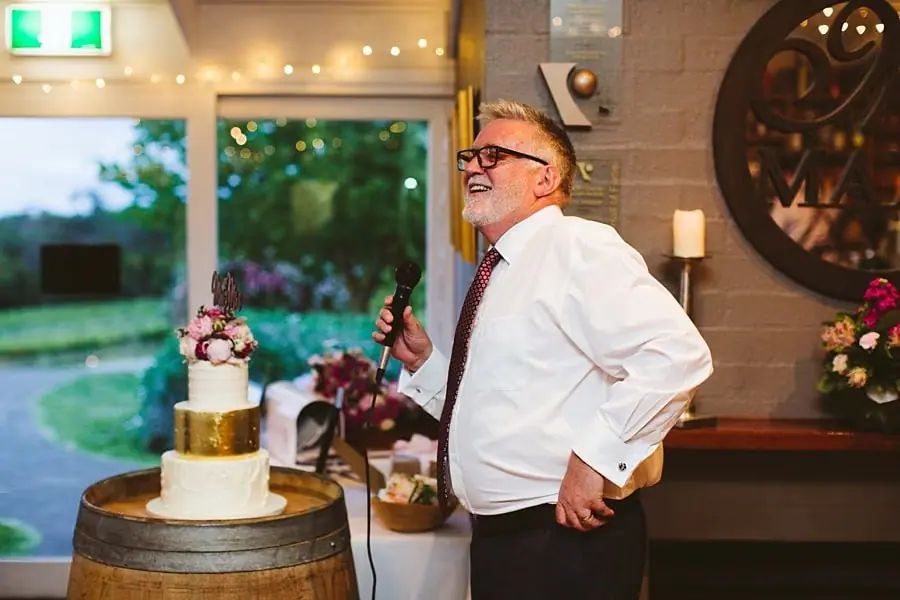 Man in glasses holding a microphone beside the wedding cake during the reception.