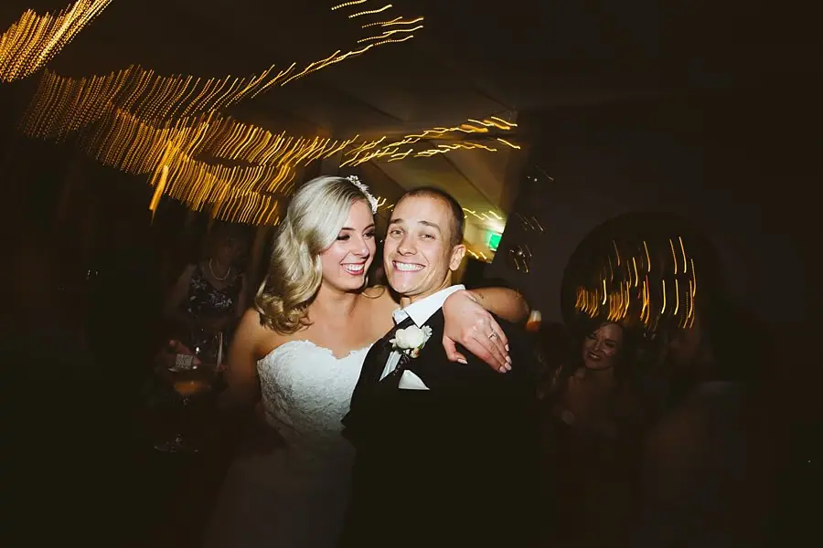 Bride and groom smiling close together beneath blurred strings of reception lights.