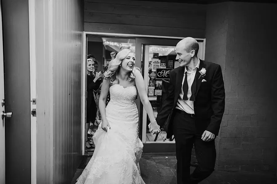 Black-and-white photograph of newlyweds smiling at each other while leaving the reception doorway.