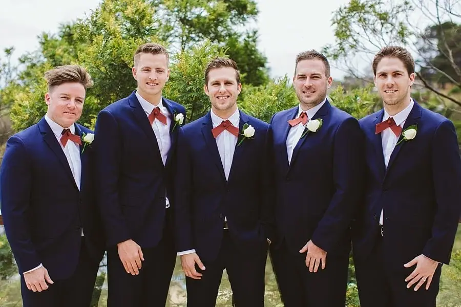 Five men in navy suits and red bow ties posing outdoors.