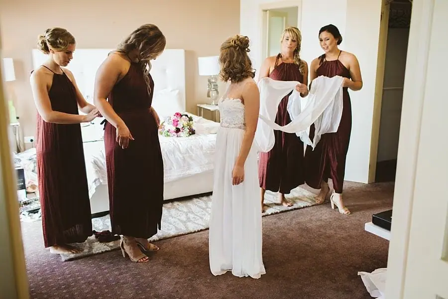 Women in burgundy dresses helping the bride get dressed.