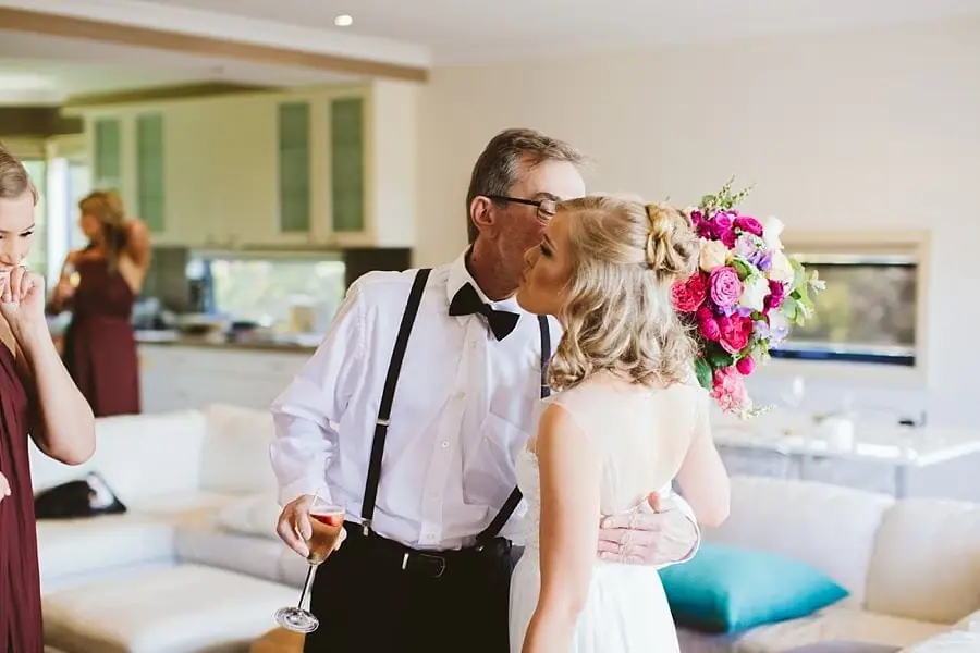 Man in braces kissing the bride's cheek indoors.