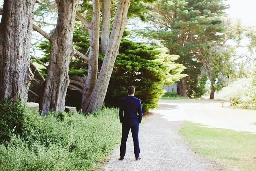 Groom standing alone on a tree-lined gravel path.