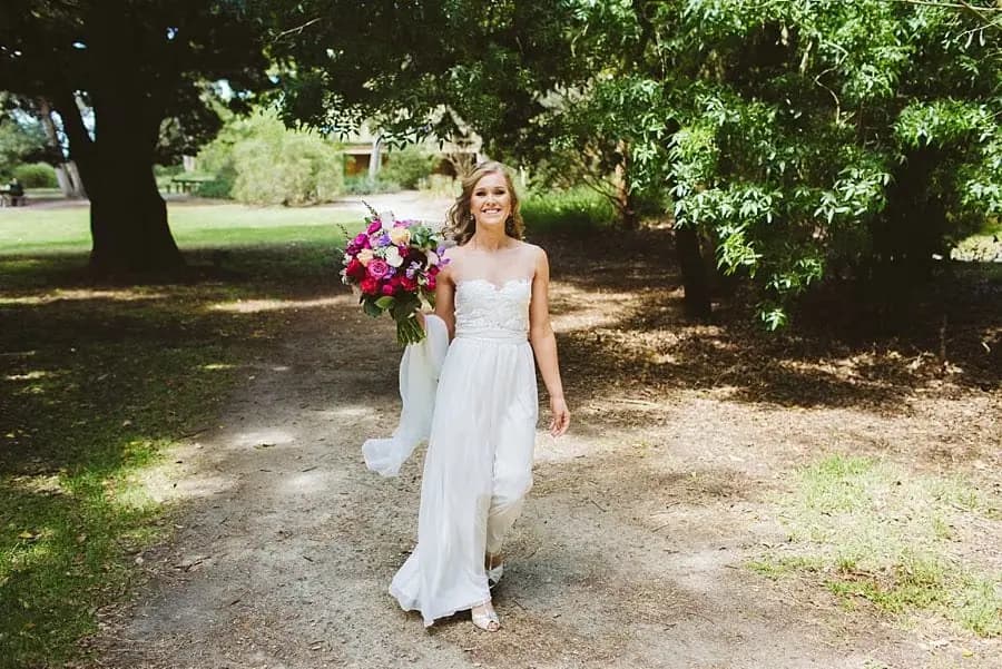 Bride walking along a shaded path carrying a colorful bouquet.