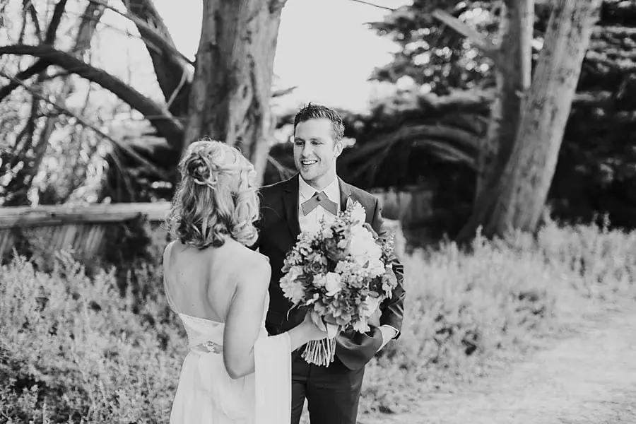 Black-and-white photograph of the groom smiling toward the bride beneath trees.