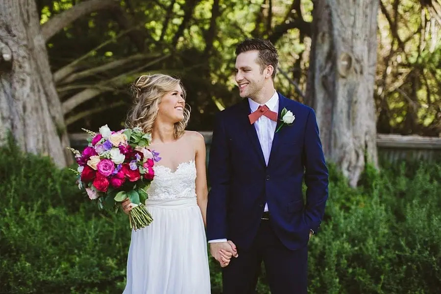 Bride and groom holding hands and smiling in a leafy garden.