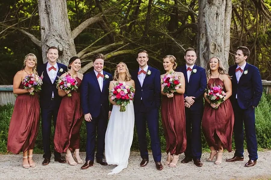 Wedding party posing in a line beneath large trees.