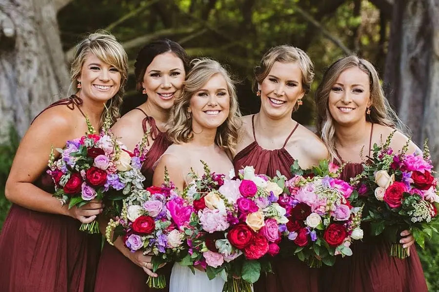 Bride and four women in burgundy dresses smiling with colorful bouquets.