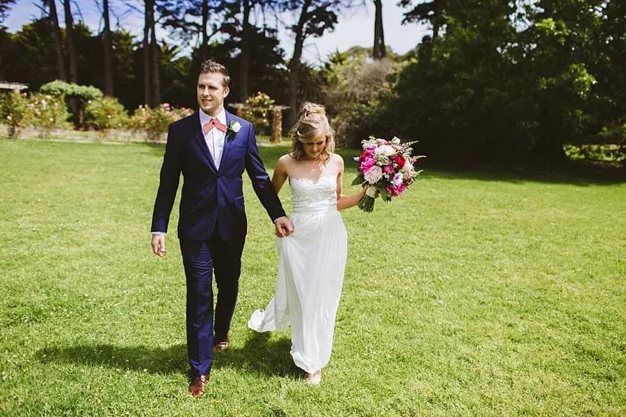 Bride and groom walking hand in hand across a green lawn.