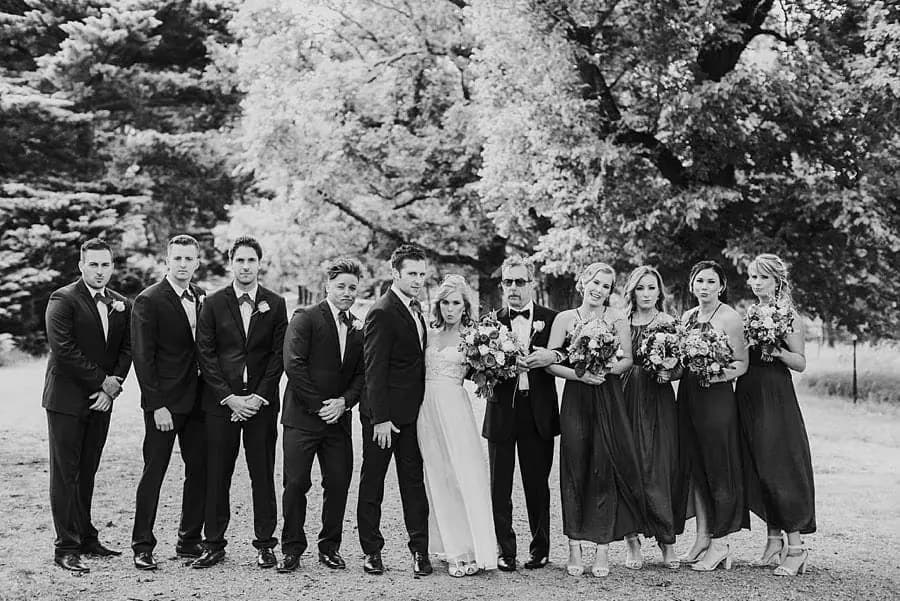 Black-and-white group portrait of the wedding party beneath trees.