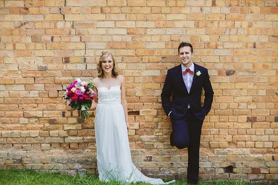 Bride and groom standing apart against a golden brick wall.
