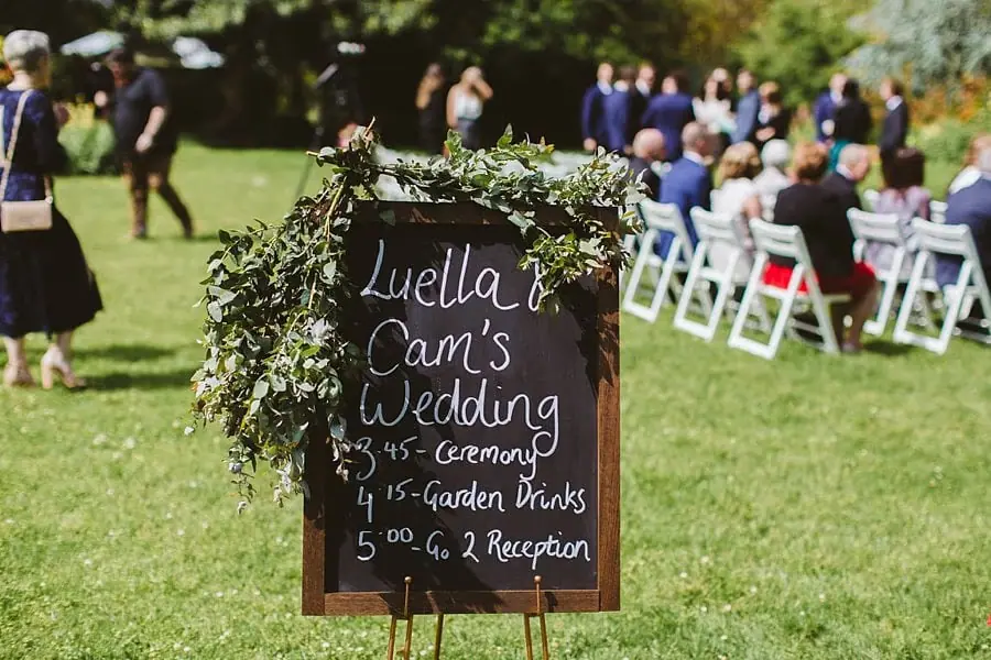 Flower-framed chalkboard displaying a wedding schedule beside outdoor ceremony seats.