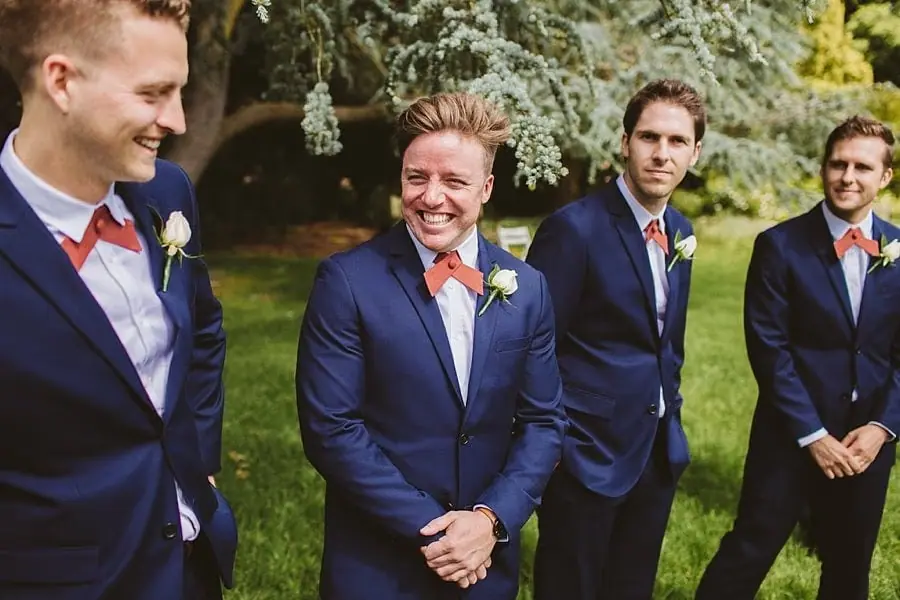 Suited men smiling together beneath leafy trees.