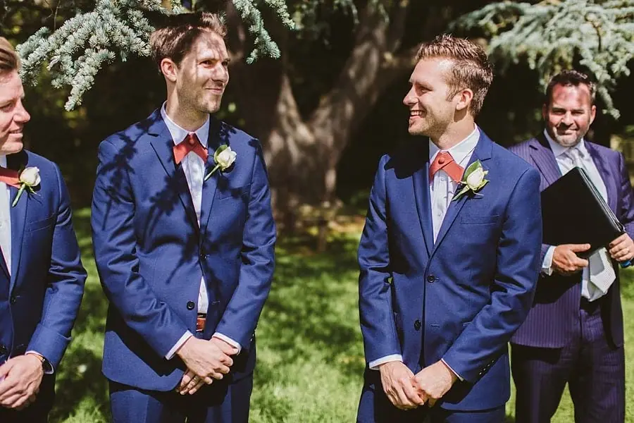 Men in navy suits and red bow ties chatting in the ceremony garden.